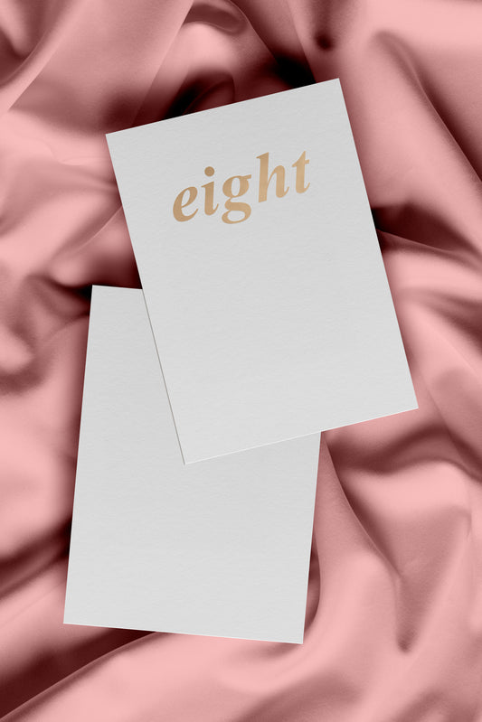 A white birthday card with 'eight' embossed in gold foil on the front. The card is laying on pink fabric background.