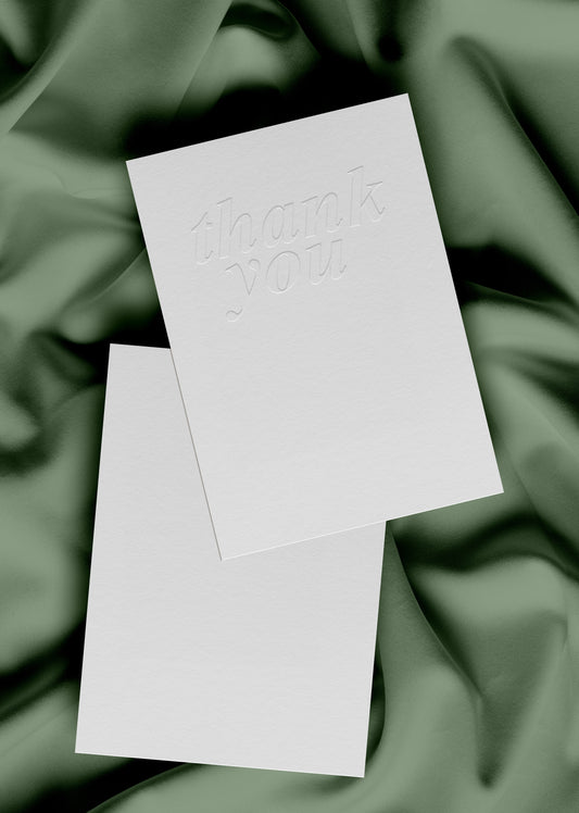 A plain white greeting card with 'thank you' embossed on the front, laying on a plain white envelope