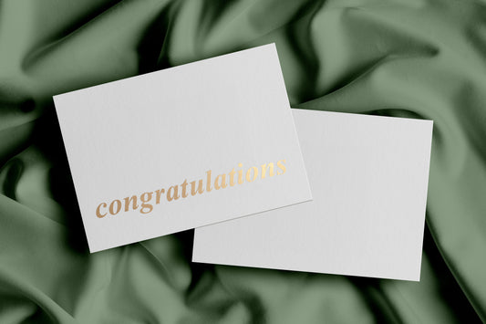 A plain white greeting card with 'congratulations' printed in gold foil, laying on a plain white envelope